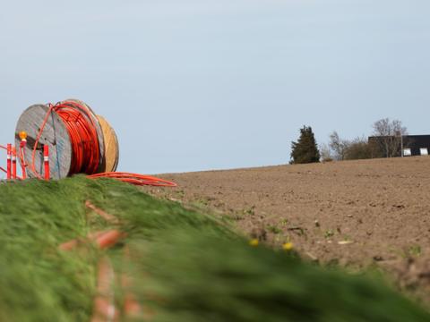 Fiberkabelrulle placeret ved grøft i en landlig mark, klar til nedgravning. Hus i baggrunden.