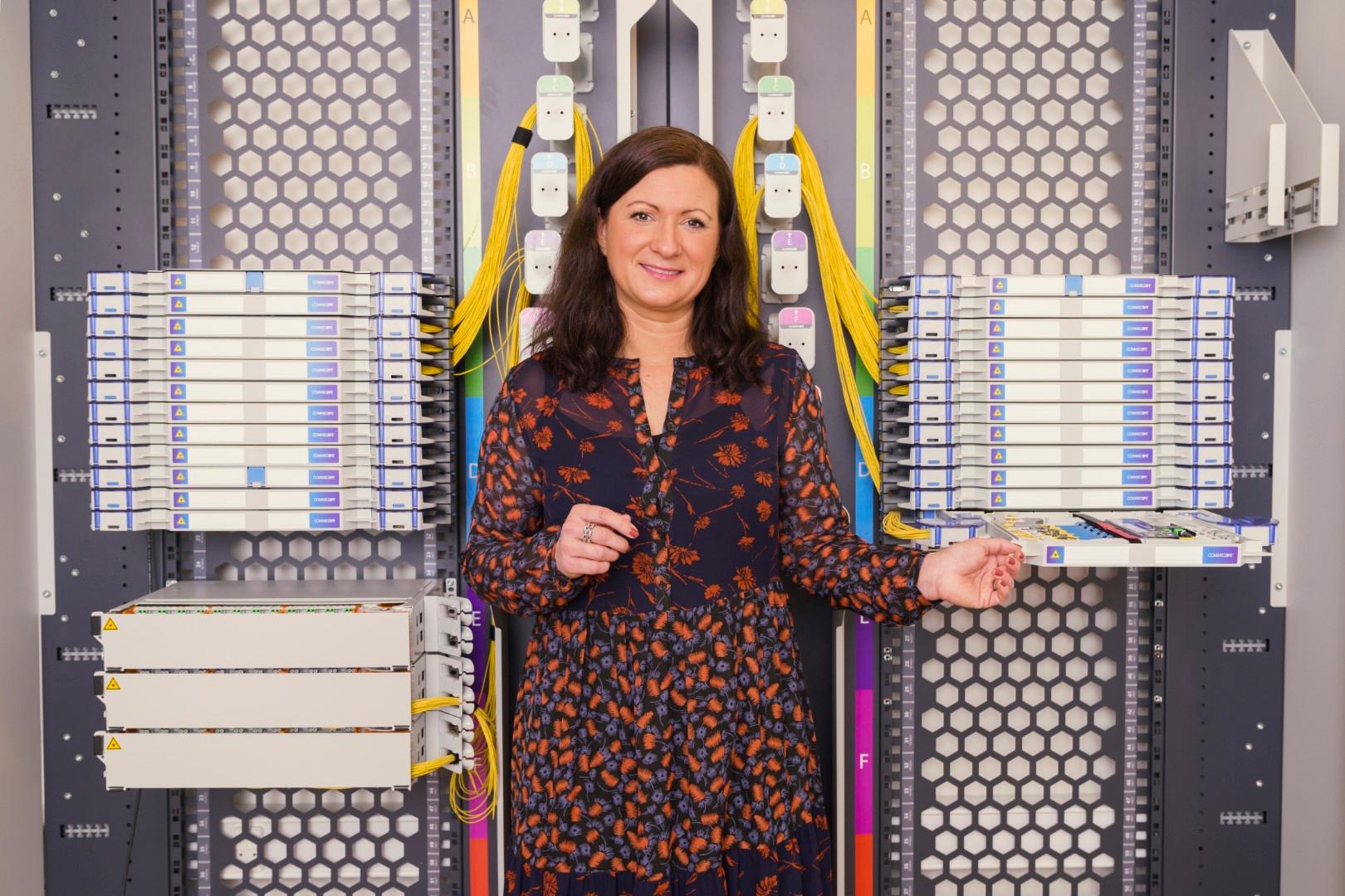 Kvinde står foran CommScope FACT-skab med organiserede patchpaneler og farvede fiberkabler. Hun smiler og holder i en af de udtrækkelige bakker, omgivet af teknisk udstyr i et moderne datamiljø.