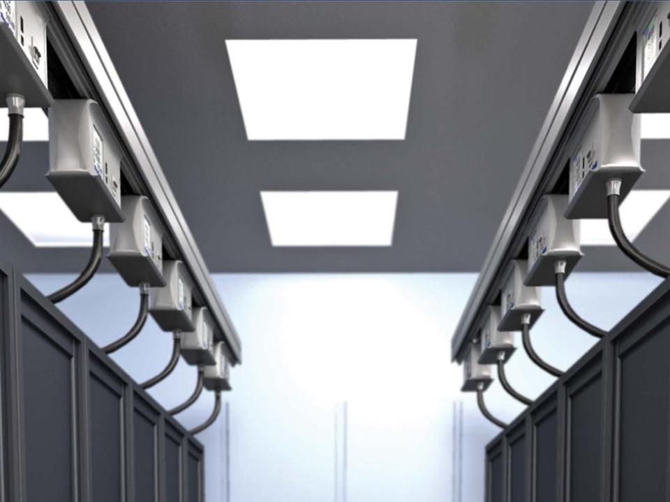 Indvendigt billede af et datacenter med sorte serverracks og loftsmonterede Starline busways eludtag, hvorfra strømkabler hænger ned til racksene. Loftet er udstyret med kraftige LED-lyspaneler.