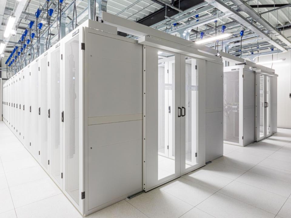 Minkels racks til datacenter. Moderne datacenter med hvide serverracks og glasdøre, opstillet i aflukkede rækker for optimal køling. Over hovedet ses kabelbakker og infrastruktur til strøm og netværk.