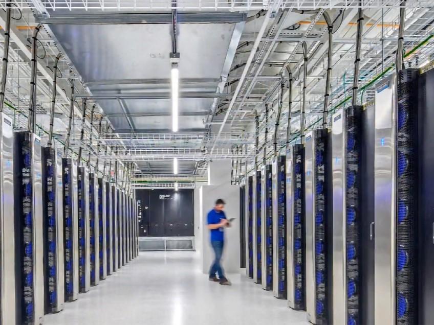 Usystems bagdørskøling hos Alcadon. Moderne datacenter med lange rækker af serverracks med bagdørskøling og en tekniker iført blåt tøj, der går gennem midtergangen med en tablet. Kabelføringer og kabelbakker er synlige i loftet.